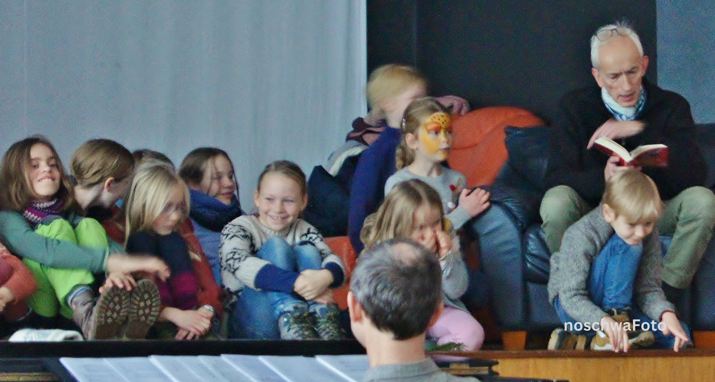Viele Kinder sitzen auf einer Bühne und lauschen gespannt einem vorlesenden Schauspieler vom Theater Lübeck